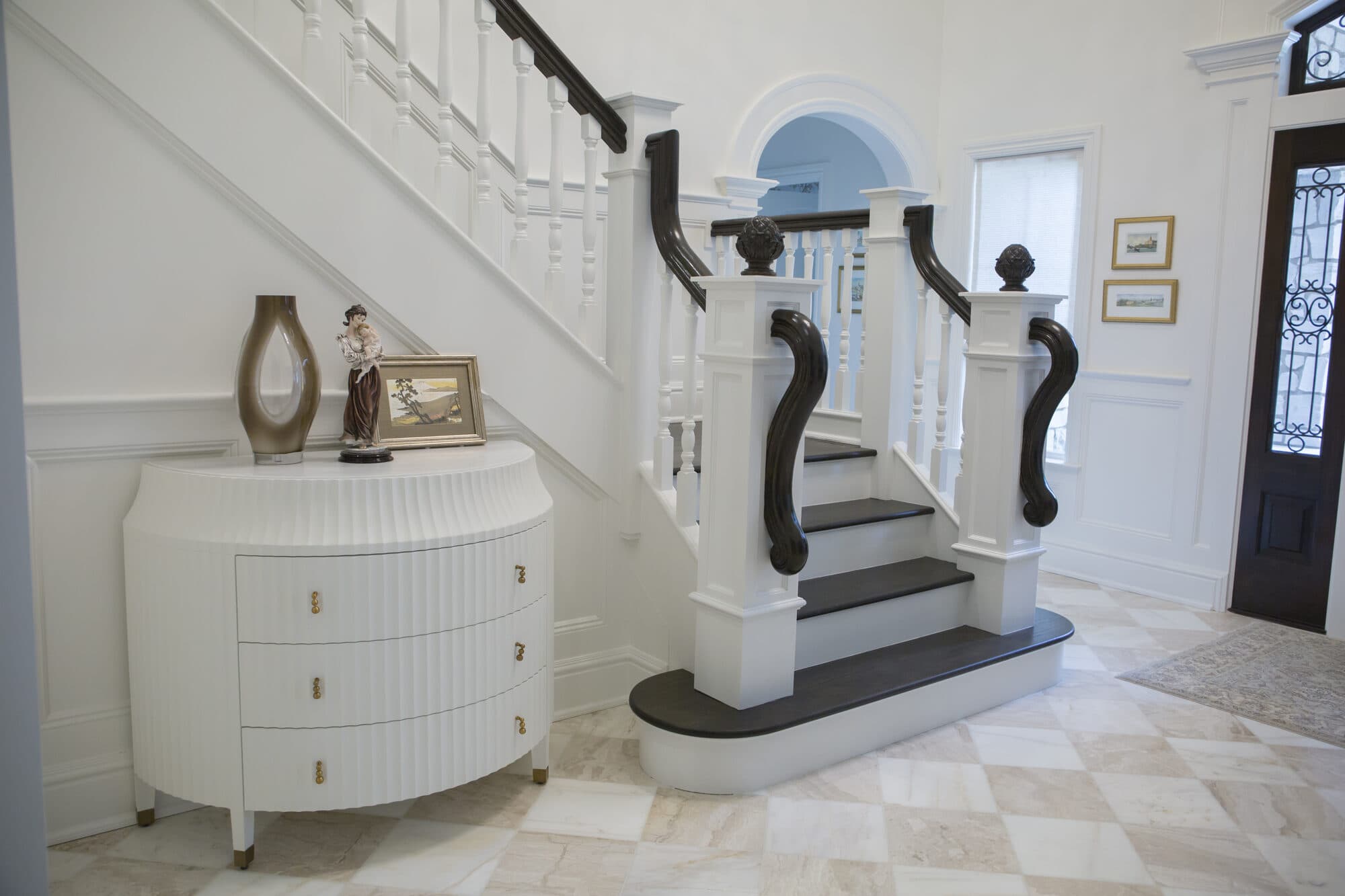 Grand white box stair with dark treads and scroll-volute newels rising from a marble-checkerboard foyer