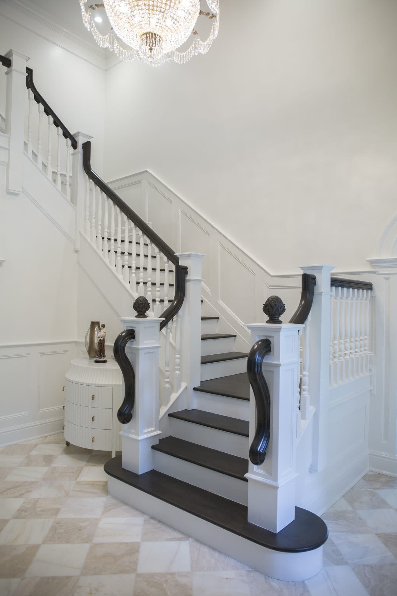 Full white two-tone box stair with dark treads, scroll-volute newels and a crystal chandelier over a marble foyer
