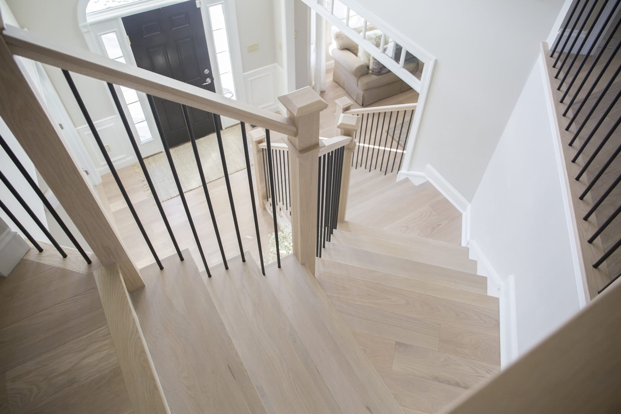 Light white-oak winder staircase with thin satin-black metal balusters descending to a foyer